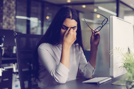 Portrait of depressed young Latin-American businesswoman sitting at computer in office holding glasses and rubbing eyes. Overworking conceptの写真素材