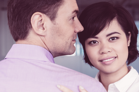 Portrait of happy Asian businesswoman embracing Caucasian male colleague or lover in office, looking at camera and smilingの写真素材