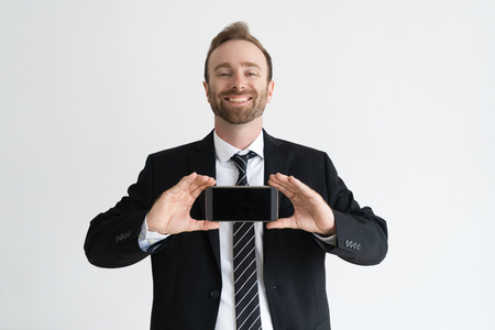 Smiling business leader holding smartphone, showing its screen and looking at camera. Promotion concept. Isolated front view on white background.の写真素材
