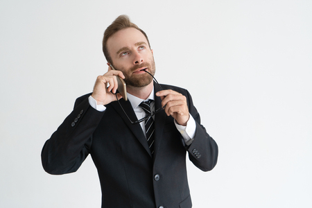 Dreamy pensive businessman having pleasant talk on smartphone. Young man in formal suit speaking on mobile phone thoughtfully and biting glasses. Communication and personal call on job conceptの写真素材