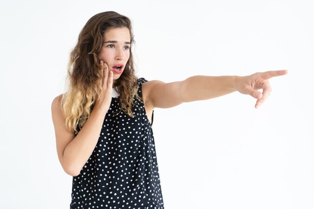 Worried pretty young woman pointing away and touching cheek. Incident concept. Isolated view on white background.の写真素材