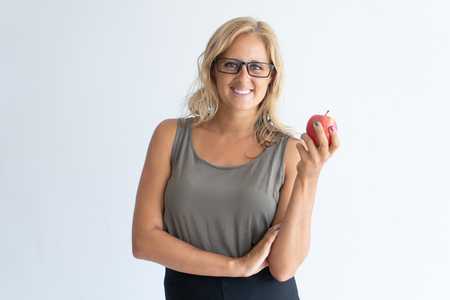 Portrait of happy mid adult businesswoman in glasses with apple. Caucasian blond-haired vegetarian wearing gray tank top looking at camera and smiling. Detox meal and healthy eating conceptの写真素材