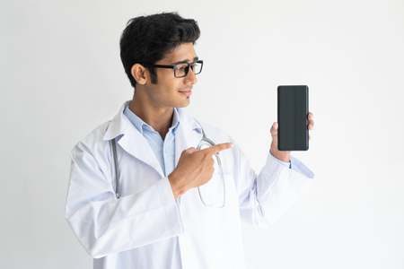 Portrait of smiling young doctor in glasses showing mobile phone. Indian medic wearing lab coat advertising medical mobile app. Mobile technology in medicine conceptの写真素材