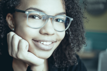 Portrait of cheerful young African American woman wearing glasses looking at camera and smiling with hand on chinの写真素材
