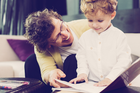 Closeup of smiling grandmother showing little grandson coloring album with blurred home interior in backgroundの写真素材