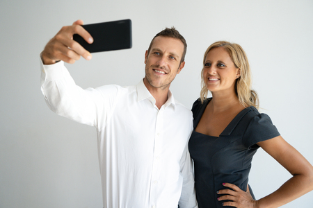 Cheerful positive young couple posing for selfie on smartphone. Happy young business couple photographing themselves for social media. Technology conceptの写真素材