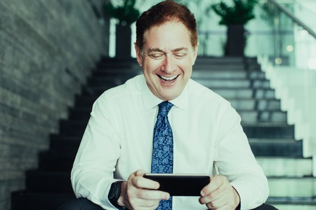 Portrait of mature Caucasian businessman wearing shirt and tie sitting on stairs, surfing internet on smartphone and smilingの写真素材