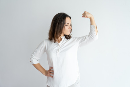 Serious proud girl showing strength gesture. Young Caucasian woman in white blouse flexing hand muscles and looking at bicep. Feminism conceptの写真素材