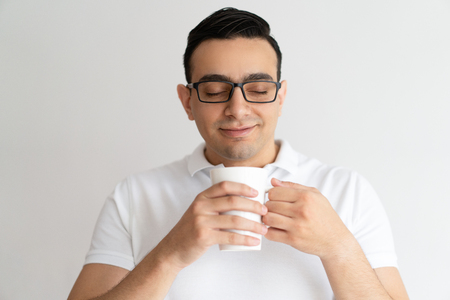 Smiling young man holding mug, smelling and drinking tea. Handsome guy enjoying drinking tea or coffee with his eyes closed. Break concept. Isolated front view on grey background.の写真素材