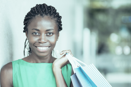 Portrait of cheerful young African American woman wearing green dress holding shopping bags, looking at camera and smilingの写真素材