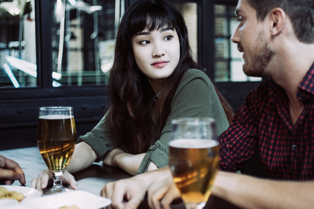 Closeup of two content young people wearing casual clothes, chatting and drinking beer at table in pub with indoor window in backgroundの写真素材