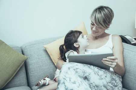 Modern mother showing educational film to daughter. Pretty girl looking at mom. They relaxing on sofa in living room. Positive family using technology. Leisure conceptの写真素材