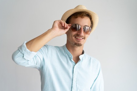 Cheerful handsome cool guy adjusting sunglasses and looking at camera. Smiling stylish young man in straw hat enjoying vacation. Summer conceptの写真素材