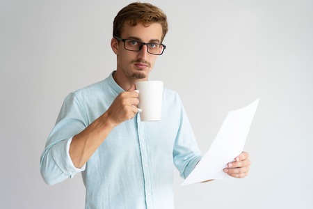 Serious busy male manager drinking coffee and reading document. Confident handsome young man in glasses analyzing paper and looking at camera. Paperwork conceptの写真素材