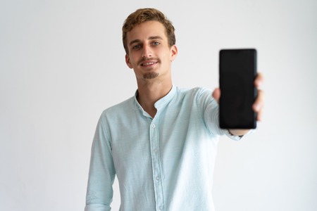 Smiling handsome young man showing smartphone to camera. Cheerful male manager sharing new mobile...