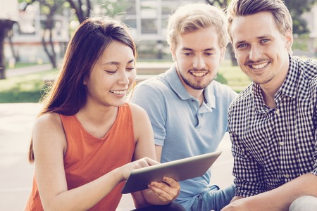 Cheerful male curator supporting students and helping them during first day in university. Young friends using digital tablet and resting in park. Education app conceptの写真素材