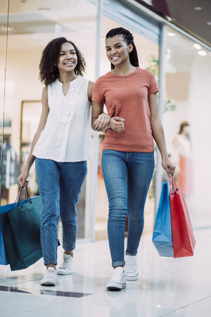 Closeup portrait of two smiling black pretty girls carrying bags, chatting and walking in shopping mall. Front view.の写真素材