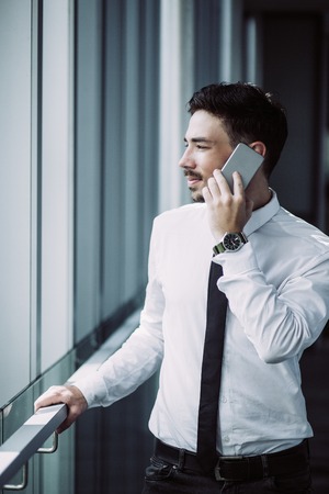 Serious young businessman having phone conversation and standing in corridor. Pensive male entrepreneur working when he being out of office. He communicating with colleague. Technology conceptの写真素材