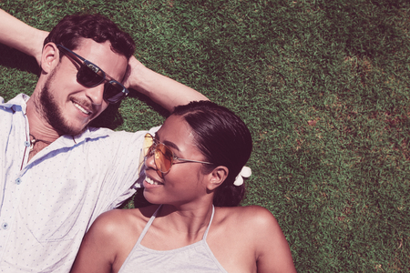 Closeup portrait of smiling young multi-ethnic couple looking at each other and lying on lawn. Top view.の写真素材