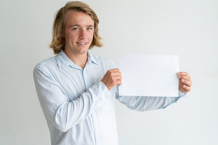 Cheerful blonde guy presenting new product. Long haired young man in white shirt holding empty sheet of paper. Presentation or promotion conceptの写真素材