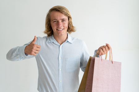 Happy young shopper satisfied with discount and purchases. Smiling Caucasian man in casual shirt holding shopping bag and showing thumb up. Buying and profit conceptの写真素材