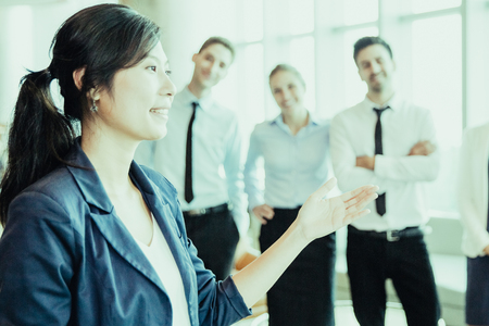 Beautiful Asian business lady holding presentation in front of her colleagues standing in background. Smiling female managing director explaining plan of work and pointing aside. Leadership conceptの写真素材