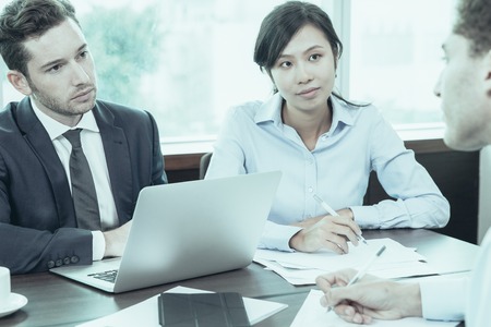 Concentrated business people listening to colleague at meeting. Serious young multi-ethnic managers discussing business strategy and sitting at table. They working as team. Job interview conceptの写真素材