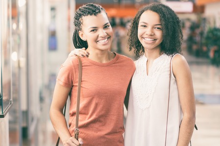 Closeup portrait of two smiling black beautiful girls looking at camera, embracing and standing in shopping mallの写真素材