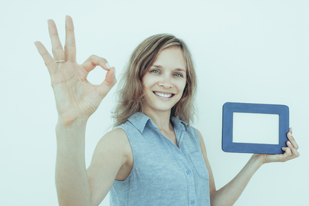Closeup portrait of smiling young attractive woman looking at camera, holding empty picture frame and showing OK sign. Isolated view on grey background.の写真素材