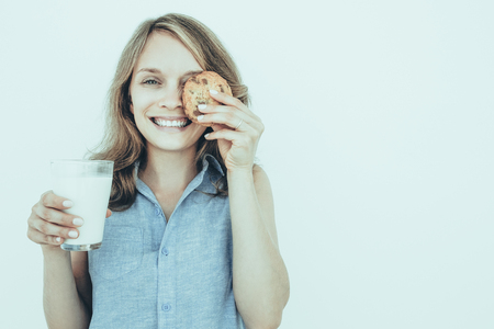 Closeup portrait of smiling young pretty woman looking at camera, holding glass of milk and covering one eye with cookie. Isolated front view on grey background.の写真素材