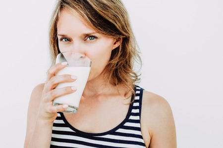 Closeup portrait of young attractive woman looking at camera and drinking milk from glass. Isolated view on grey background.の写真素材