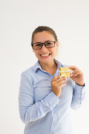 Smiling overjoyed office employee happy about her gift. Young Caucasian woman in formal wear showing small gold gift box at camera. Presenting jewelry conceptの写真素材