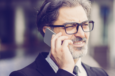 Closeup portrait of smiling middle-aged handsome business man looking away and calling on smartphone outdoorsの写真素材