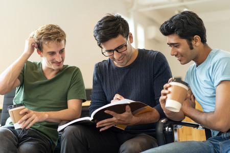 Cheerful students learning notes in workbook and analyzing information together. Positive young men thinking of task and sitting on chairs while drinking coffee. Task solution conceptの写真素材