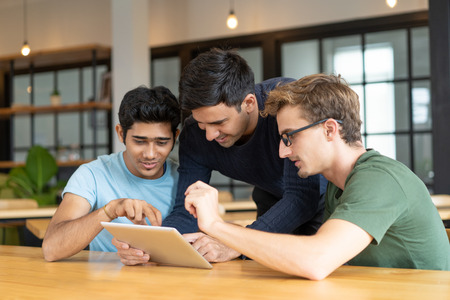 Positive developers discussing their startup during break. Three cheerful young men sharing tablet,...