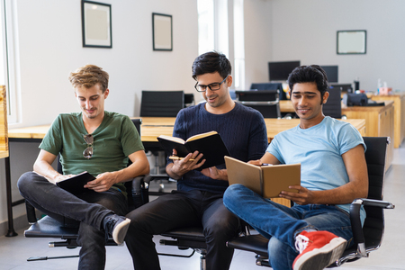 Three smiling fellow students studying and reading textbooks. Young men sitting in armchairs in classroom or library. Education concept. Front view.の写真素材