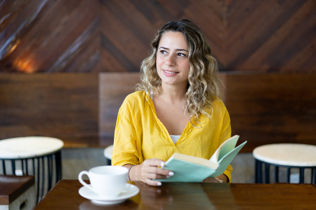 Dreamy smiling beautiful student with book sitting at table. Pensive young woman studying in cafe. Education conceptの写真素材