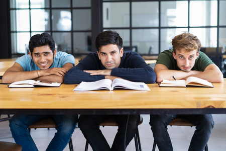 Three content students lying on desk and looking at camera. Young men sitting and studying in classroom or library. Education and rest concept. Front view.の写真素材