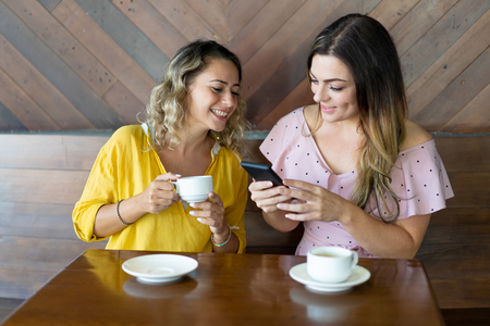 Girls drinking coffee and discussing new mobile application. Smiling attractive woman showing photo on smartphone screen to friend. Meeting with friend conceptの写真素材
