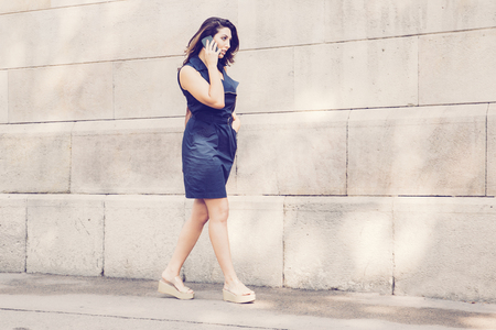 Serious young Latin-American woman wearing blue dress walking along wall and talking on cellphoneの写真素材