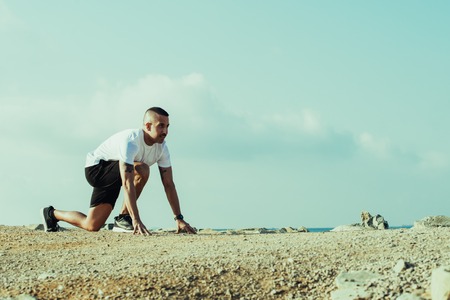Aspirated sporty man in starting position before race. Concentrated young sportsman leaning on ground and waiting for start. Intent on winning conceptの写真素材