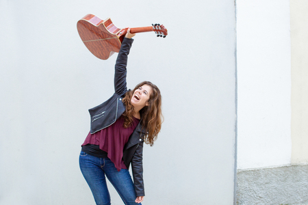 Ecstatic female guitar player celebrating success. Excited long haired woman in jeans and leather jacket raising guitar overhead. Positive emotions conceptの写真素材