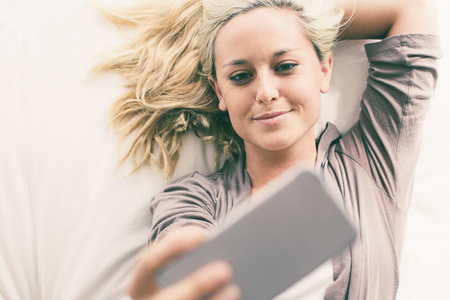 Portrait of young Caucasian woman lying in bed, taking selfie with smartphone and smilingの写真素材
