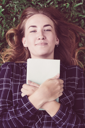 Closeup portrait of content young beautiful woman looking at camera, embracing book and lying on grass in park. Top view.の写真素材