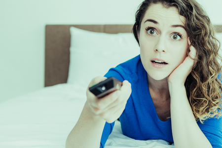 Closeup portrait of surprised young beautiful woman using remote control, watching TV and lying on bed in bedroom. Front view.の写真素材