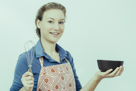 Happy young confectioner preparing icing for cake and looking at camera. Cheerful young housewife in apron holding bowl and whisk opposite white background. Cooking conceptの写真素材