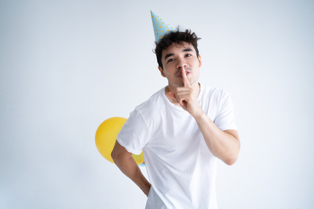 Cheerful guy in birthday hat preparing surprise party. Joyful young mixed race man hiding balloons behind back and showing shh gesture. Birthday surprise and secret conceptの写真素材