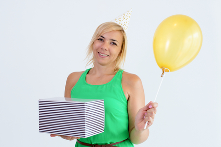 Smiling pretty woman holding balloon and gift box. Positive lady wearing party hat and looking at camera. Birthday party and gift concept. Isolated front view on white background.の写真素材