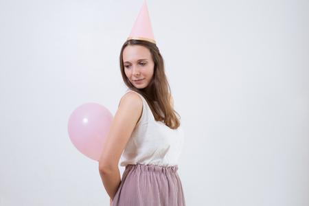 Dreamy girl in pink party hat holding balloon behind back and looking down. Calm pretty young woman lost in thoughts. Birthday alone conceptの写真素材