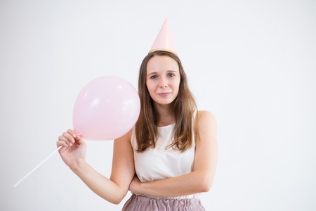 Content girl holding pink balloon and looking at camera. Attractive young woman in party cap standing against white wall. Celebration event conceptの写真素材